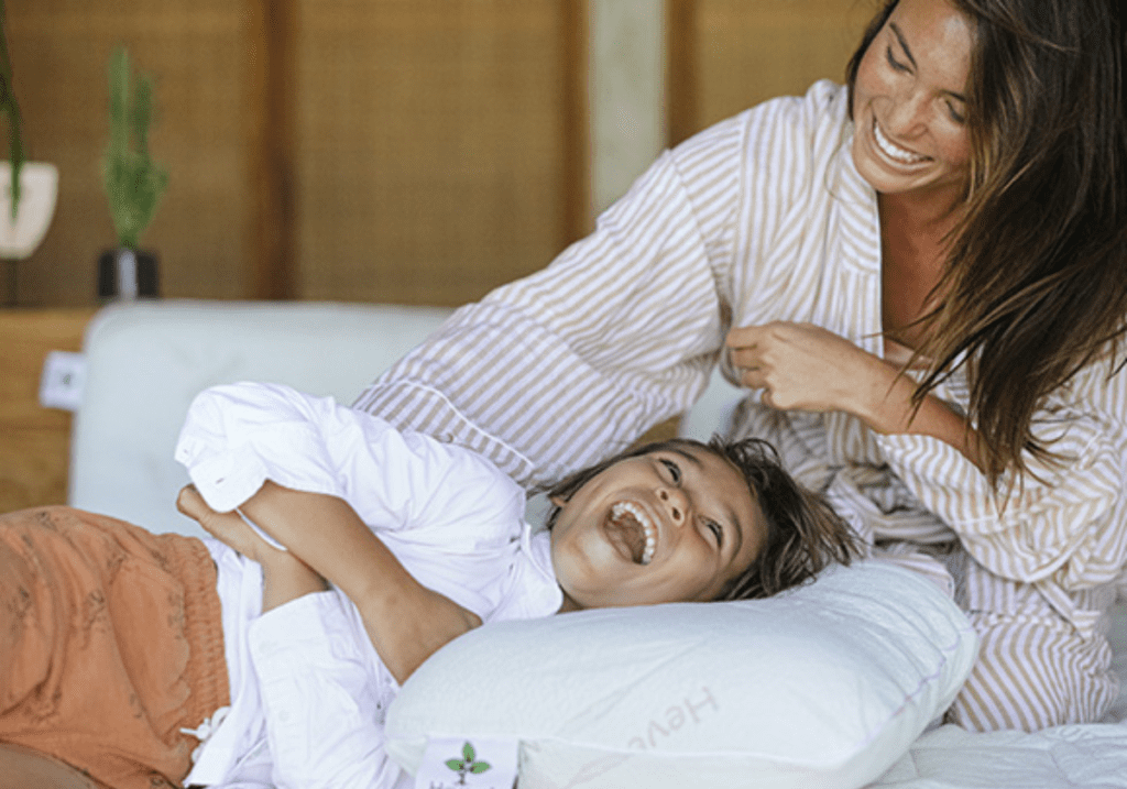Mom and child enjoying playtime on a bed featuring a Heveya® Natural Organic Latex Pillow. The pillow's natural anti-microbial and dust mite resistant properties promote a healthier sleep environment.
